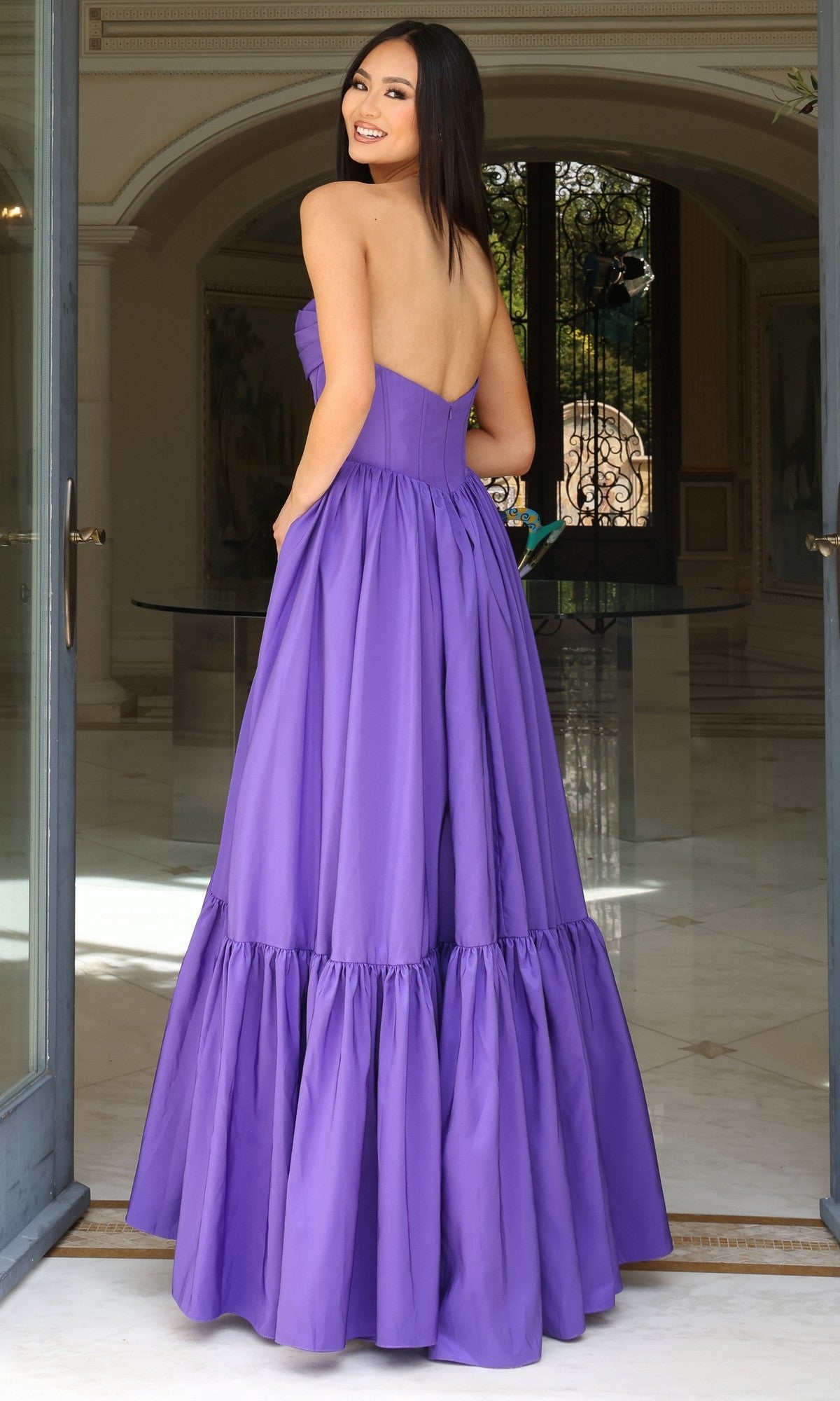 A woman stands indoors before ornate glass doors, wearing the Jovani 47883— a strapless long ruffled prom dress with a sweetheart neckline and corset-style bodice, glancing back over her shoulder.